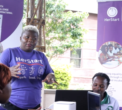 A woman in a HerStart shirt speaks as others listen during an outdoor group meeting with laptops and banners.